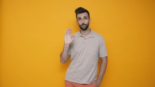 Happy brunet man wearing t-shirt showing goodbye gesture at the camera in the yellow studio