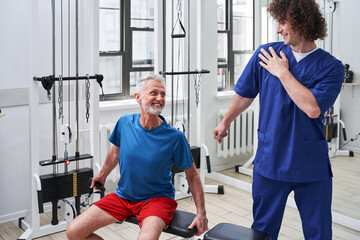 Fototapeta na wymiar Cheerful young trainer smiling and explaining something to his senior patient