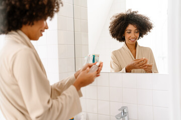 African american woman smiling and holding skin tonic in bathroom