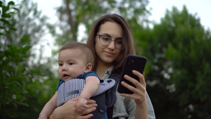 A young woman with a child stands with a phone in her hands in the park. Mom in glasses with her son in her arms. Newborn son in a kangaroo-backpack. Close-up.