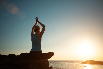 A woman does yoga, meditating in the lotus position on the ocean during sunset.
