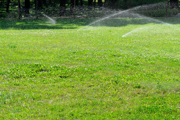 Close-up automatic garden watering system with different sprinklers installed under turf. Landscape design with lawn hills and fruit garden irrigated with smart autonomous sprayers at sunset time.