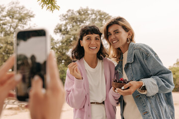 Woman is holding smartphone, taking photo of two smiling young caucasian girls spending time outdoors. Brunette and blonde wear casual clothes. Mood, lifestyle, concept.