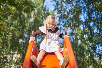 little girl schoolgirl plays in uniform in the yard. The child rolls from the children's slide on the playground.