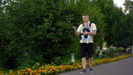 A young dad walks with a child sitting in a kangaroo backpack. A single father walks through the park with a phone in his hands.