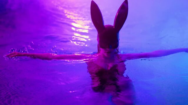 Sensual woman in leather BDSM mask moving seductively in swimming pool water under neon color light. Halloween party, attractive chick masquerade bunny enjoying nighttime games for adults