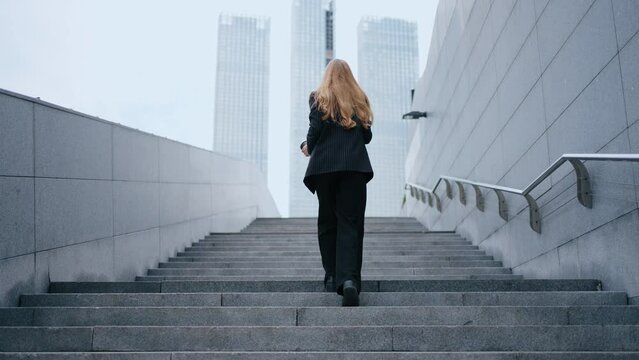 View from the back of a business woman climbing stairs in a modern city against the backdrop of city buildings. The camera follows the woman climbing the career ladder