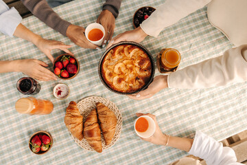 Top view of cinnamon apple pie, croissants, drinks are arranged on picnic table. Four pairs of female hands cover lunch. Concept of delicious food