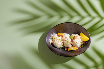three scoops of ice cream with nuts and peach slices on a dark plate on a green background with a shadow from a palm tree, top view space for text