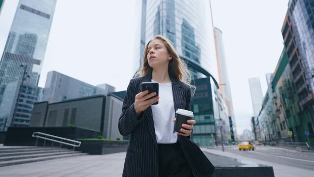 successful businesswoman is walking in city in morning, using smartphone and drinking coffee