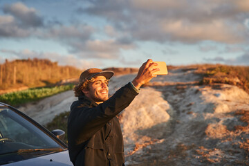 Young man sitting at his car at the beach and making selfie at smartphone