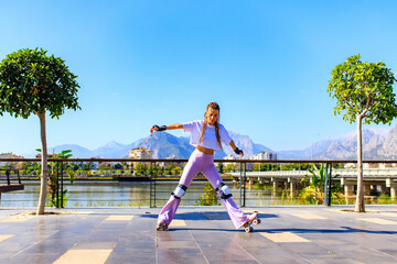 Obraz premium Portrait of beautiful blonde teenager girl with cool pigtails , white t-shirt and flared pink pants skating on rollers in park