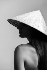 Fashion concept. Black and white portrait of beautiful woman with naked shoulder and long dark hairs wearing traditional cone-shaped Asian cane hat and cover her face in shade. Face is in focus