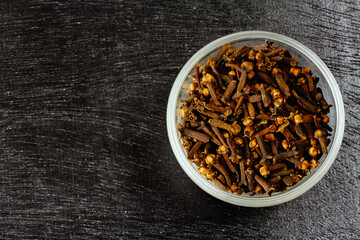 Cloves in glass bowl on black wooden background, top view, copy space. Herbal cloves have a fragrant and spicy flavor.