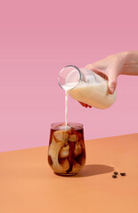 Ice coffee in a tall glass with cream poured over and coffee beans. A woman's hand pours cream into...