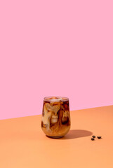 Ice coffee in a tall glass with cream poured over and coffee beans on pink background.