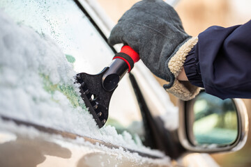 Hand in glove holding a snow scraper and removing ice from a car window with rear view mirror in background. Concept of danger on roads in winter season. Maintenance of vehicle in cold temperatures. - obrazy, fototapety, plakaty