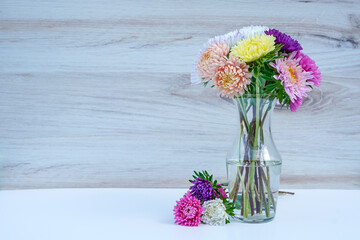 Glass vase full of colorful aster flowers with several laying on the table. Space for text.