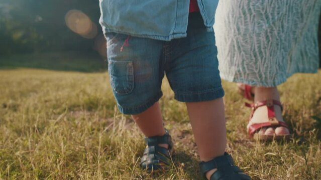 Child learns to walk baby takes first steps with the mother, helping the baby to teach the child in nature. Small legs walk on the grass, close-up. Child learns to walk in the park. Baby first steps.