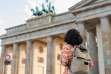 back view of curly tourist with backpack standing near brandenburg gate in berlin.