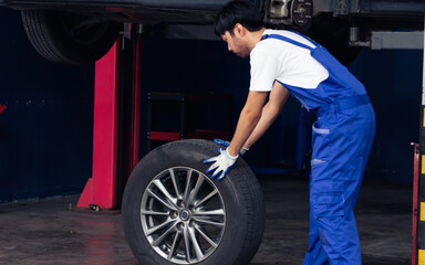 Naklejka na ściany i meble Asian handsome male mechanic wearing uniform, changing rubber tyre or tire wheel, working, reparing in garage at car or automobile maintenance service center or shop. Industry Concept.