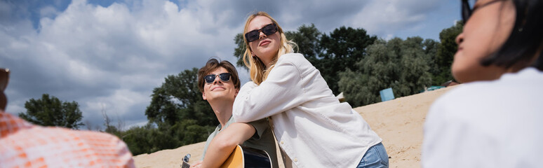 smiling man in sunglasses playing guitar near blonde woman and interracial friends, banner.