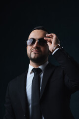 Portrait of a modern man in a fashionable suit, a man with a beard straightens his sunglasses, a man in a black suit on a dark background