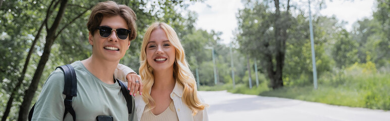 young and happy travelers smiling at camera in countryside on summer day, banner.