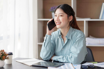 Smiling beautiful young Asian business woman enjoying phone call with customer and commenting at the office while working on a laptop computer.