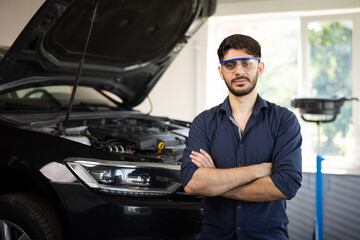 Portrait Bearded Handsome Car Mechanic is Posing in a Car Service. He Wears a Jeans Shirt and Safety Glasses. His Arms are Crossed. Specialist Manager Looks at a Camera and Smiles