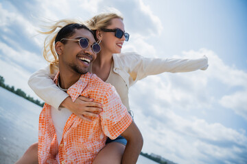 happy african american man in sunglasses piggybacking excited girlfriend on summer day.