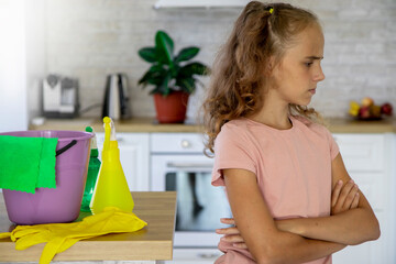 Selective focus on a blonde girl child who shows signs with her hands that she does not want to clean the house. There are detergents, a bucket and rubber gloves on the table