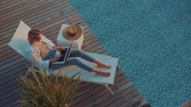Top down view of a young entrepreneur woman with blonde hair is working on her computer in by the resort pool in summer time on a sunbed at sunset. Remote working anywhere, the concept of a digital no