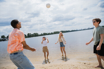 joyful multiethnic friends playing beach volleyball near river on summer day.