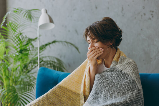 Young Sick Ill Sad Woman Wears White Wrapped In Plaid Cough Cover Mouth With Hand Sit On Blue Sofa Couch Stay At Home Hotel Flat Spend Time In Living Room Indoors Grey Wall. People Healthcare Concept.