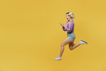 Full body side view young blonde woman 20s wear pink tied shirt white t-shirt jump high hold in hand use mobile cell phone run fast isolated on plain yellow background studio People lifestyle concept