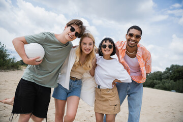 cheerful multiethnic friends in sunglasses smiling at camera on beach.