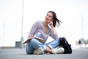 Young woman sitting outside with pen and book looking away