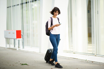 Woman walking with suitcase bag while using mobile phone in city
