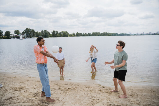 interracial men playing beach volleyball near women standing in water.