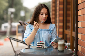 young woman eating cake at outdoors cafe and read message on her smart phone.