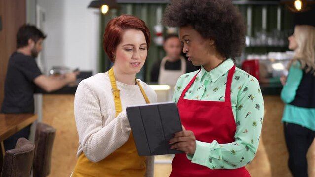 Female employee explaining job to new staff colleague. African American worker holding tablet discussing work standing inside coffee shop small business