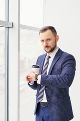 Fototapeta premium A young man in a business suit looks at his watch with a cup of coffee. A businessman in a shirt and tie and a dark jacket. A bearded man in a cafe or restaurant.