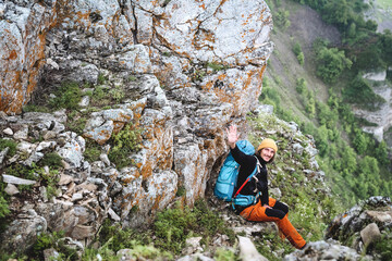 Obraz premium A man sits on the edge of a cliff looking into the camera, a guy with a backpack on a hike smiling waving his hand, greeting people in nature, mountain tourism.