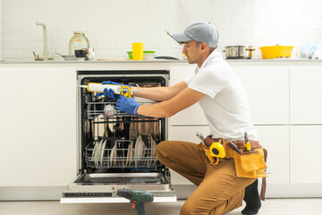 Repairman in uniform repairs dishwasher in kitchen. Young man specialist unscrews parts with screwdriver checking state of trays for dish side view.