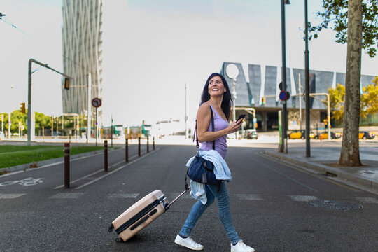 Happy woman looking away while walking in city with luggage and mobile phone
