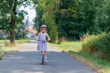 Portrait of active little preschool girl with glasses and helmet riding scooter on road in park outdoors on summer day. Seasonal child activity sport. Healthy childhood lifestyle