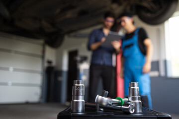 Male Mechanic Talking to Manager Under Vehicle in Car Service. Specialist is Showing Info on a Tablet Computer. Empowering Man in Modern Workshop