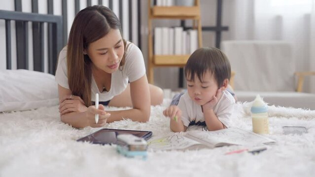 Asian mother play with her little boy on bed with boy paint using color pencil paint on color book and woman use tablet to show the example color. A bottle of milk also put beside of the boy.