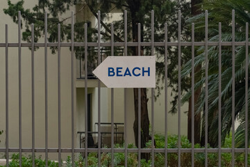 A white sign indicating the direction towards the beach against the background of a metal fence. A sign of the direction to the sandy beach in the resort town.
